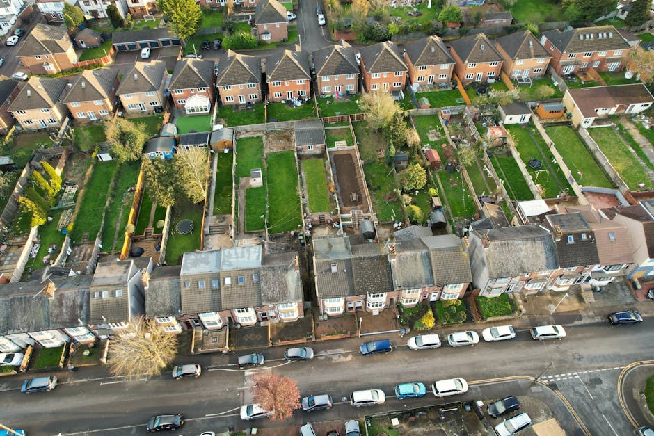 An aerial view of a residential area showing a row of terraced houses with small front gardens and parked cars along the street. Behind the houses, the back gardens are visible, featuring a variety of